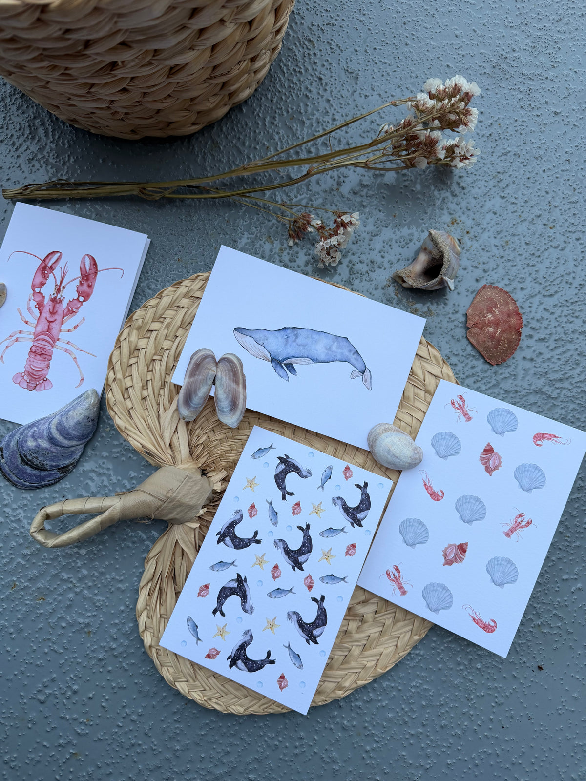 A styled flat-lay of four greeting cards featuring nautical illustrations, arranged on a woven mat with seashells, dried flowers, and a wicker basket on a textured gray surface.
