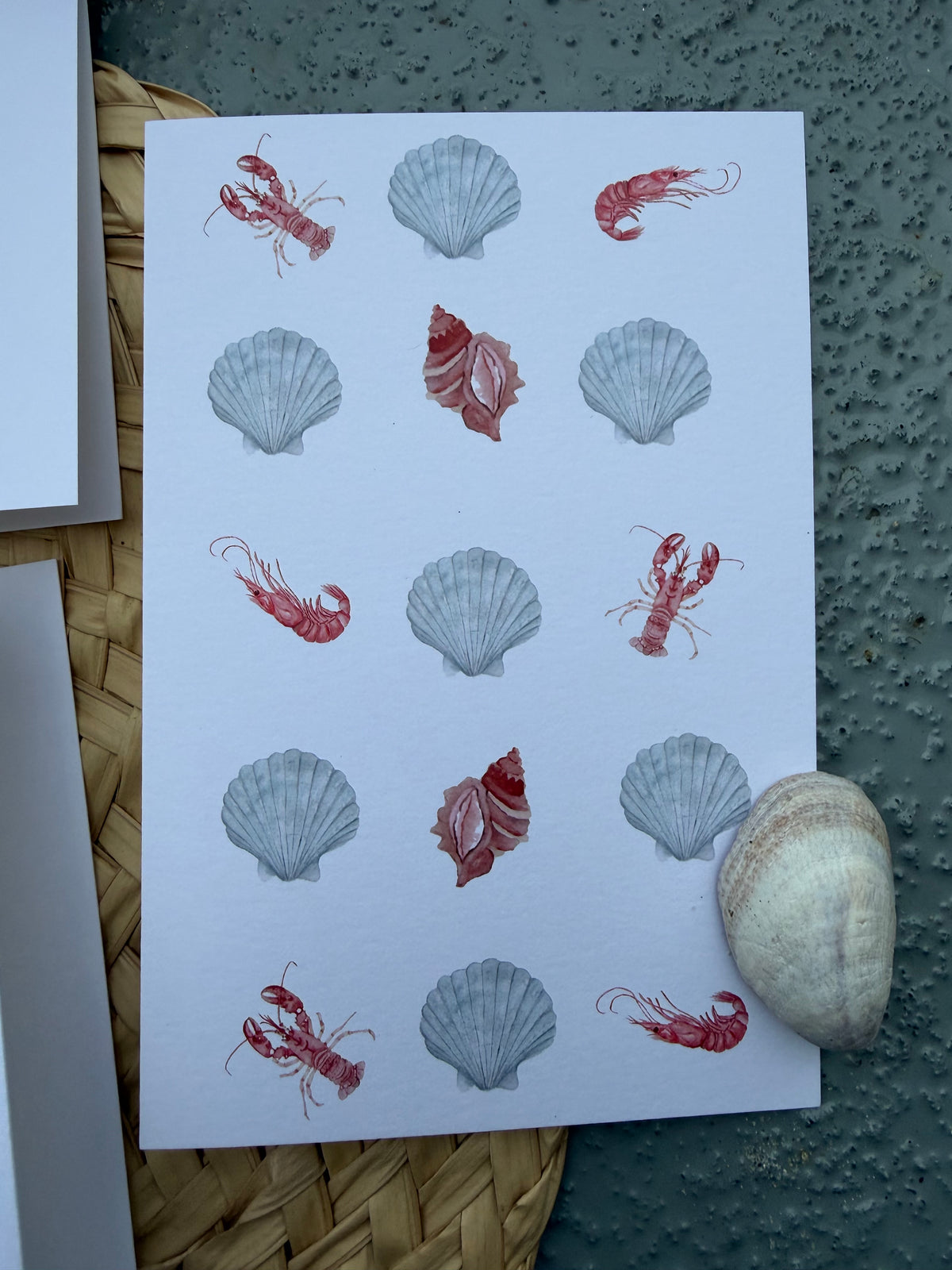 A greeting card featuring watercolor illustrations of red lobsters and gray seashells, set on a woven wicker mat, with a real seashell resting in the corner, against a dark, textured background.