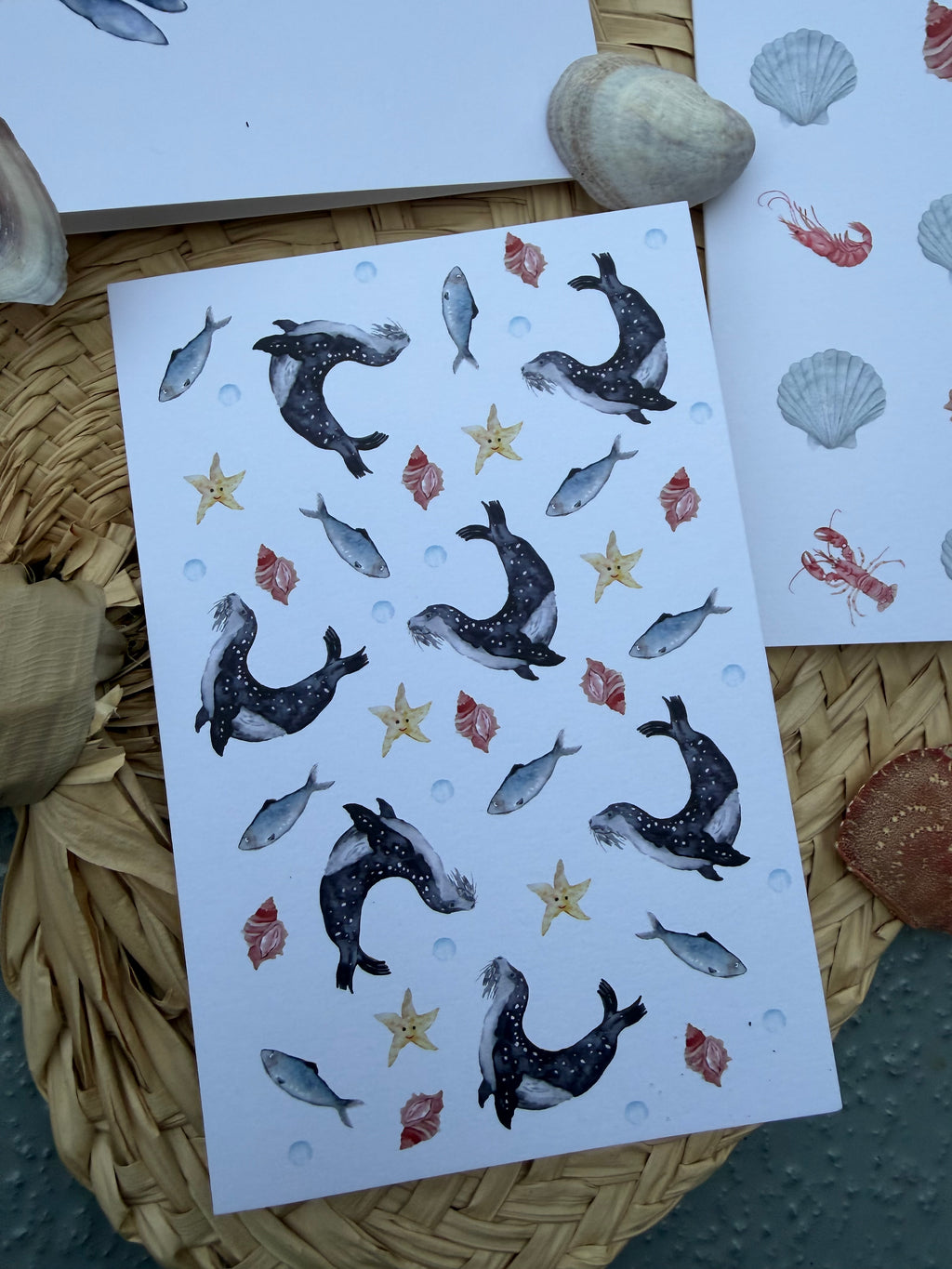Greeting card featuring sea-life illustrations, including seals, fish, starfish, and seashells, displayed on a woven mat, with seashells used as props.