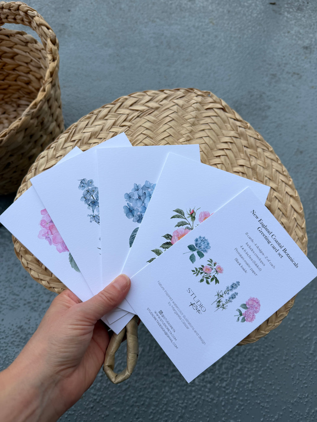 A hand holds a fanned-out set of white printed cards featuring watercolor floral illustrations in pink, blue, and green, placed on top of a round woven straw basket against a gray textured background.