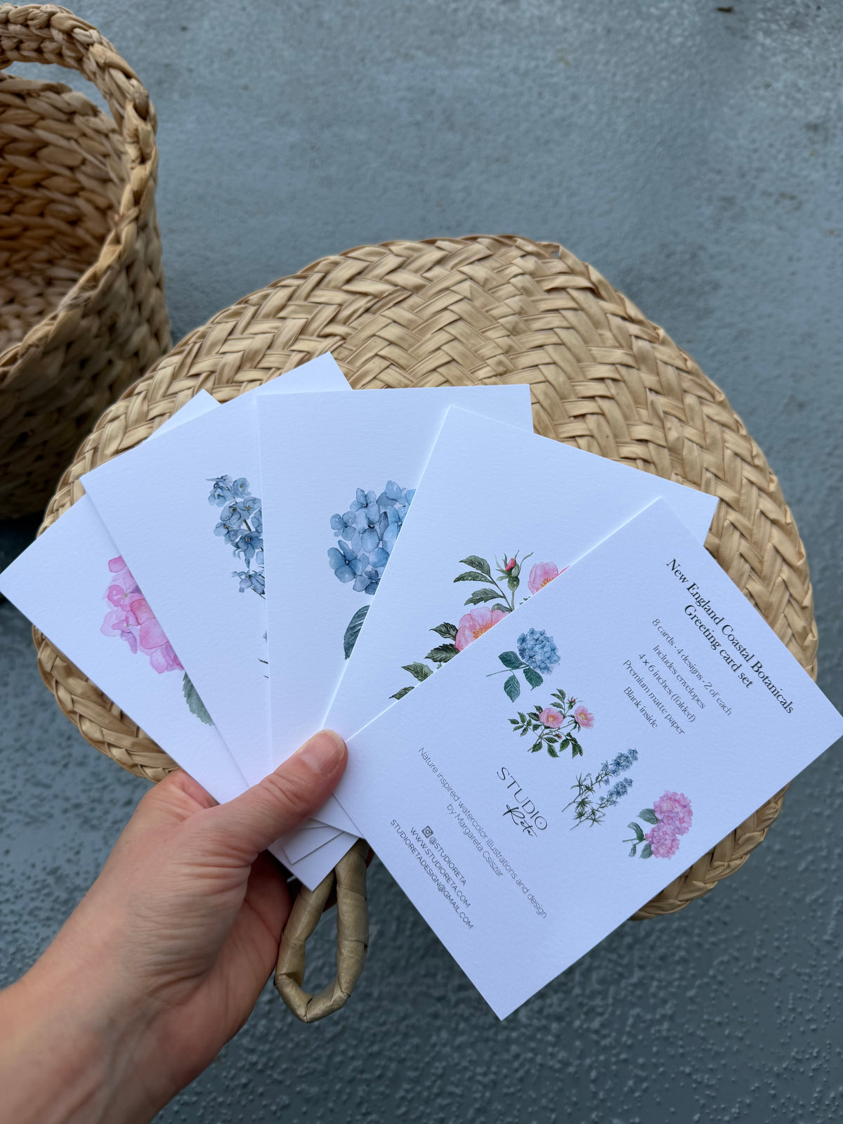 A hand holds a fanned-out set of white printed cards featuring watercolor floral illustrations in pink, blue, and green, placed on top of a round woven straw basket against a gray textured background.