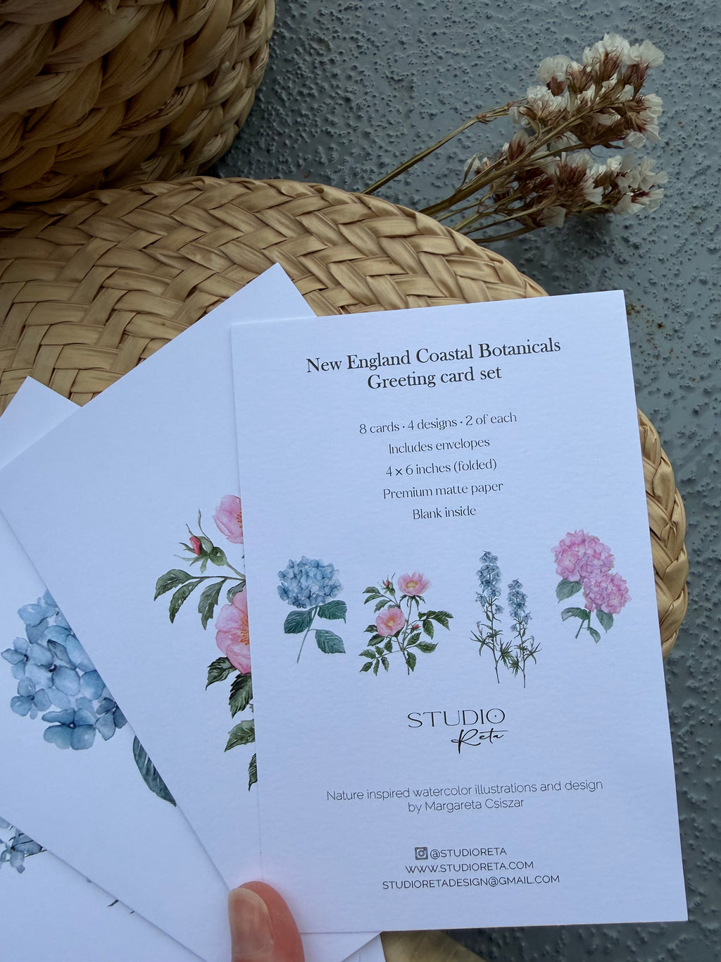 A hand holds a stack of botanical greeting cards featuring watercolor illustrations of flowers, placed on woven wicker mats against a textured gray wall.