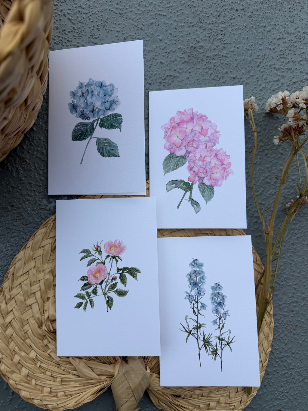 Four botanical greeting cards featuring blue and pink hydrangeas, delphinium and beach roses, arranged on a woven wicker tray against a textured gray wall.