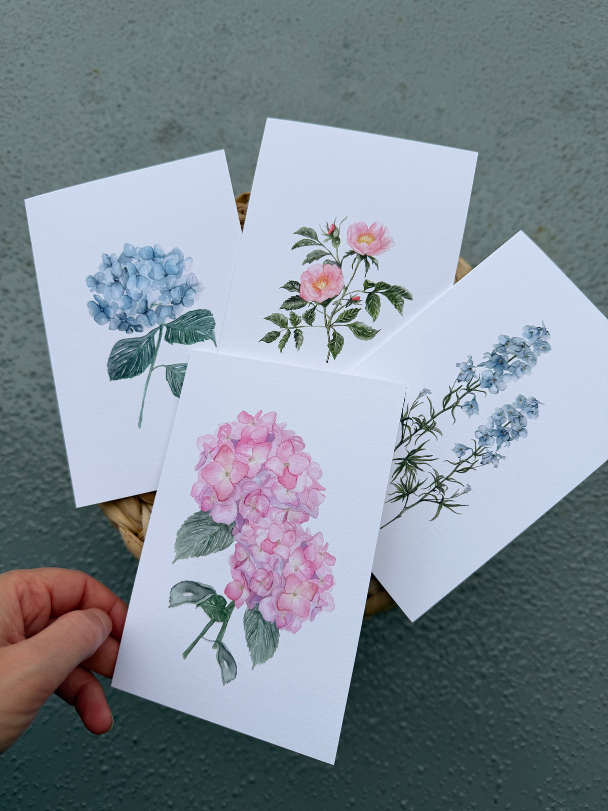 Four botanical greeting cards with watercolor-style flowers are fanned out on a textured green surface, with a hand holding the front print.